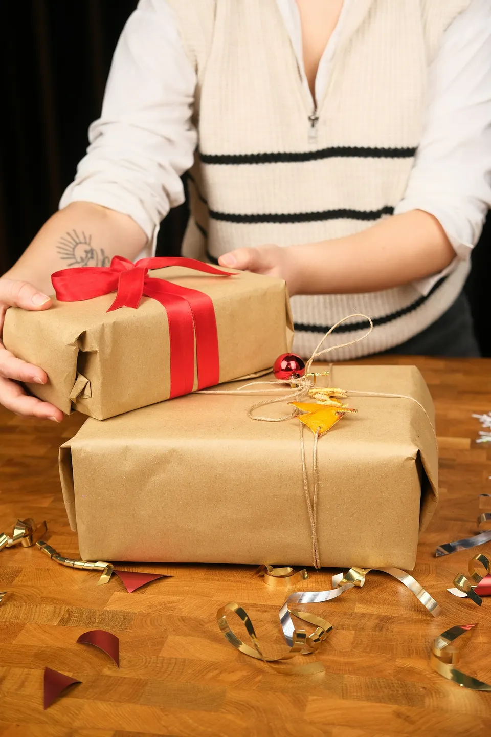 Hands wrapping gifts in brown kraft paper, one tied with a red satin ribbon